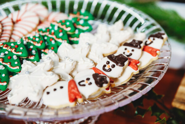 The Season of Giving at Catered by Design 3 Snowmen and Christmas tree-shaped cookies on display on a clear plate.